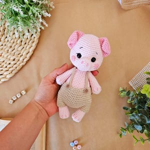May include: A pink crocheted pig wearing brown overalls with buttons. The pig has black eyes and a pink snout.