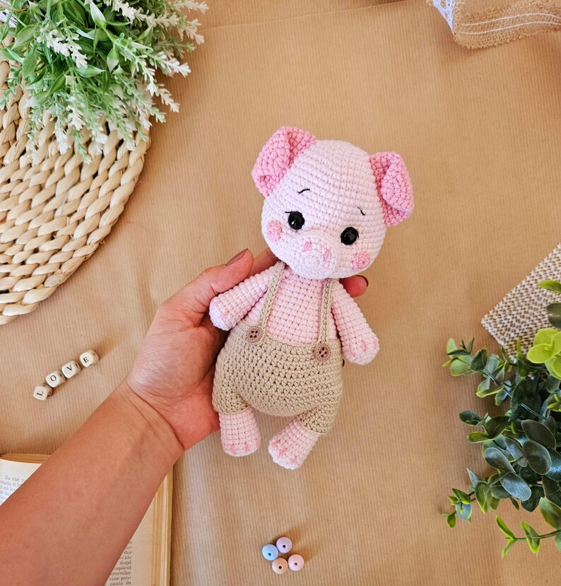 May include: A pink crocheted pig wearing brown overalls with buttons. The pig has black eyes and a pink snout.