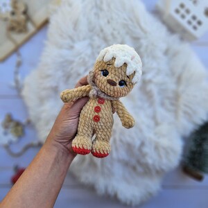 May include: A crocheted gingerbread man doll with a white icing-like head, a tan body, and red feet. The doll has a small scarf, black eyes, and red button details. It is a handmade item.