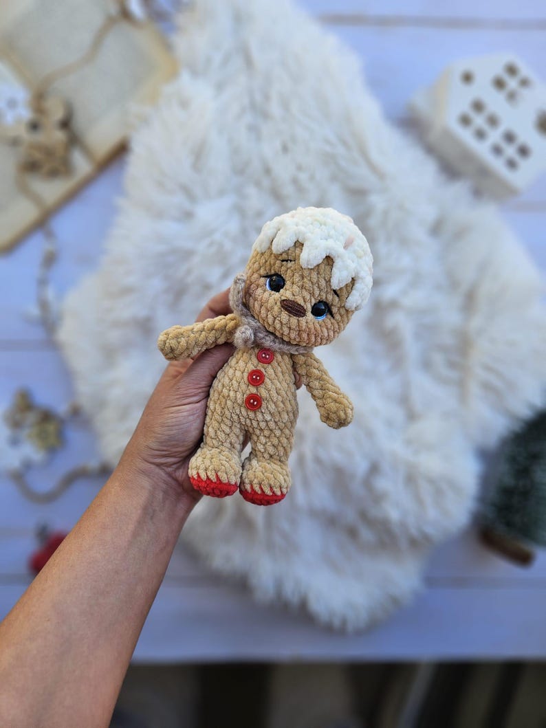May include: A crocheted gingerbread man doll with a white icing-like head, a tan body, and red feet. The doll has a small scarf, black eyes, and red button details. It is a handmade item.
