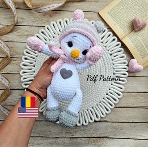 May include: A handmade white snowman plushie with a gray heart, wearing a pink and gray hat, scarf, and mittens. The snowman has a yellow carrot nose and black eyes. The text "Pdf Pattern" is visible on the surface.