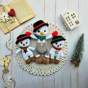 May include: Three handmade crochet snowmen with black hats, red accents, and carrot noses. The snowmen are wearing scarves and mittens, and are arranged on a decorative circular mat. A small Christmas tree and a decorative house are also in the image.