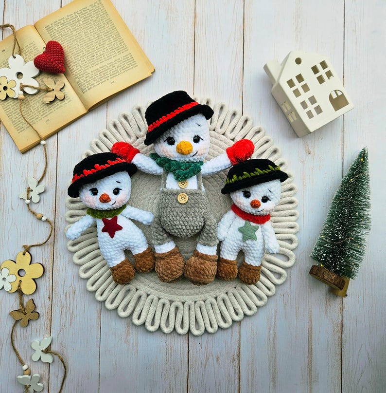 May include: Three handmade crochet snowmen with black hats, red accents, and carrot noses. The snowmen are wearing scarves and mittens, and are arranged on a decorative circular mat. A small Christmas tree and a decorative house are also in the image.