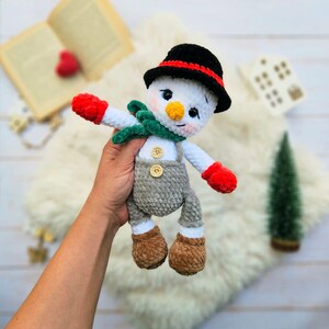 May include: A handmade snowman doll with a black hat, red and green scarf, and gray overalls. The snowman has a carrot nose, rosy cheeks, and red mittens. The doll is made of soft, textured yarn and is a festive winter decoration.