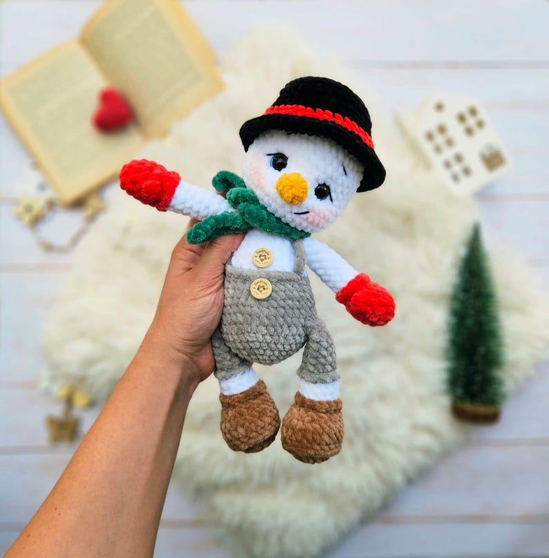 May include: A handmade snowman doll with a black hat, red and green scarf, and gray overalls. The snowman has a carrot nose, rosy cheeks, and red mittens. The doll is made of soft, textured yarn and is a festive winter decoration.