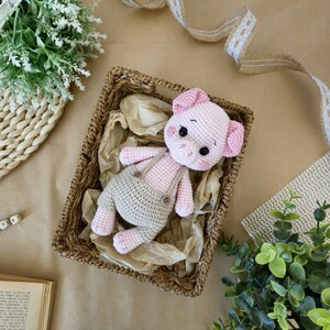 May include: A pink crocheted pig wearing overalls sits in a wicker basket. The pig has a brown nose and black eyes. The basket is filled with brown paper.