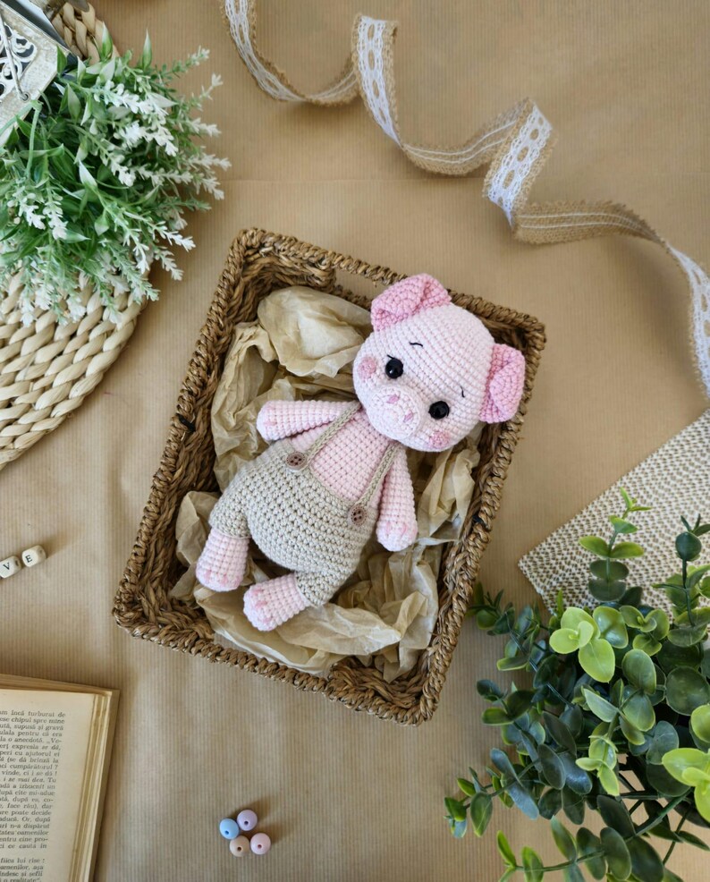 May include: A pink crocheted pig wearing overalls sits in a wicker basket. The pig has a brown nose and black eyes. The basket is filled with brown paper.