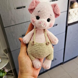 May include: A crocheted pink pig wearing brown overalls with two buttons. The pig has black eyes and a pink nose.
