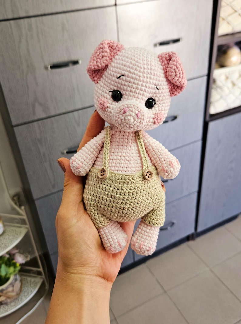 May include: A crocheted pink pig wearing brown overalls with two buttons. The pig has black eyes and a pink nose.