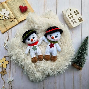 May include: Two handmade crochet snowmen with black hats, red and green scarves, and star accents. The snowmen have carrot noses and brown boots. They are displayed on a fluffy white surface with a small Christmas tree and decorative elements.
