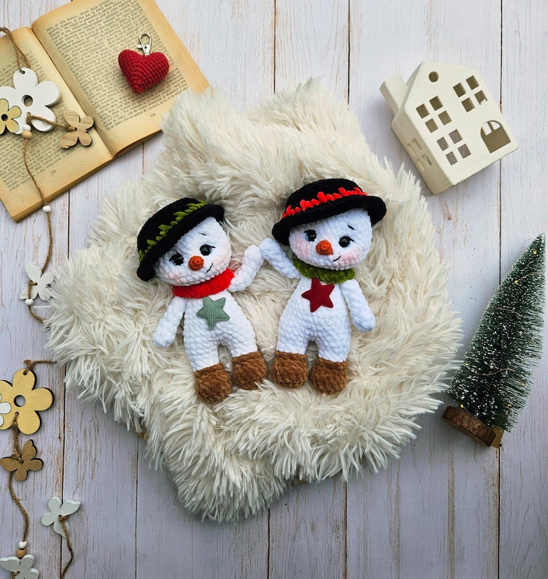 May include: Two handmade crochet snowmen with black hats, red and green scarves, and star accents. The snowmen have carrot noses and brown boots. They are displayed on a fluffy white surface with a small Christmas tree and decorative elements.