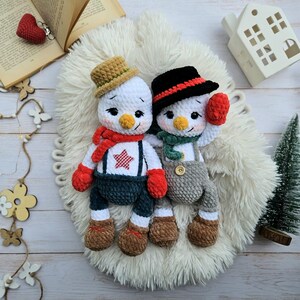 May include: Two handmade crochet snowmen dolls. One snowman wears a brown hat, red scarf, and blue overalls with a red star. The other snowman has a black hat, green scarf, and gray overalls. Both have carrot noses and red mittens.