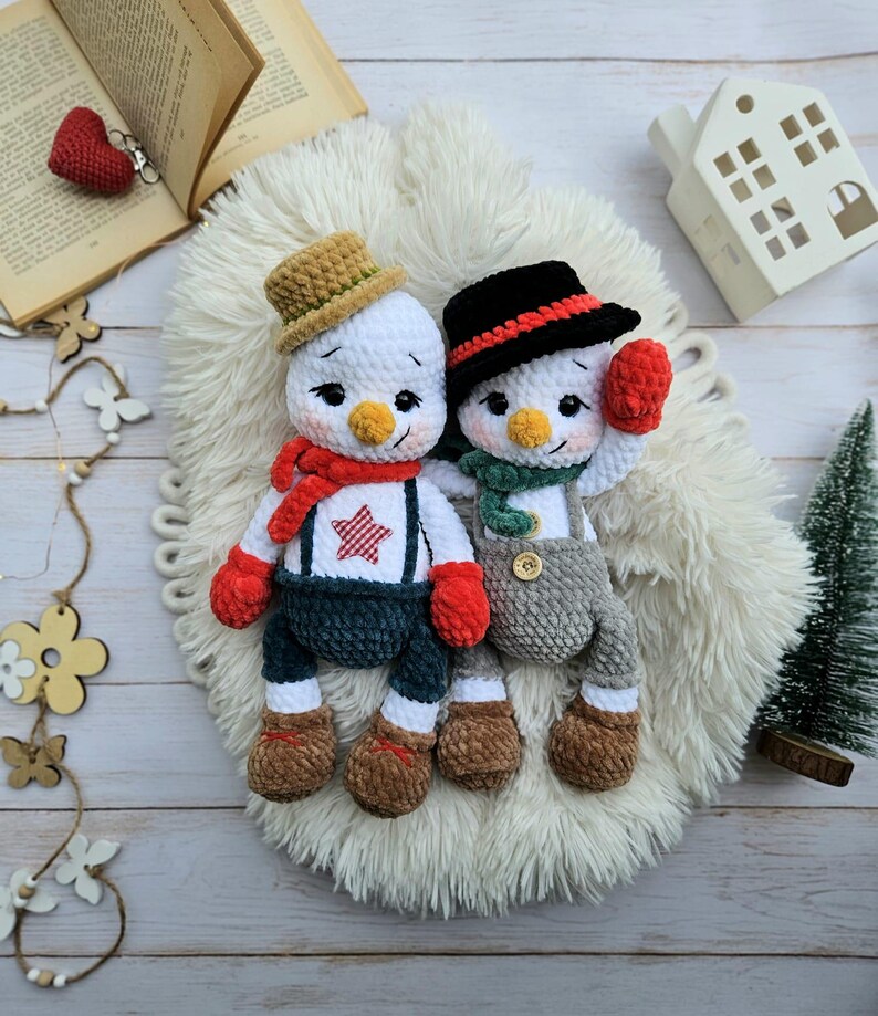 May include: Two handmade crochet snowmen dolls. One snowman wears a brown hat, red scarf, and blue overalls with a red star. The other snowman has a black hat, green scarf, and gray overalls. Both have carrot noses and red mittens.
