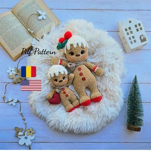 May include: Two crocheted gingerbread man dolls, one large and one small, rest on a fluffy white surface. The larger doll has a scarf, red buttons, and a decorative hat. The smaller doll holds a red heart. The text "Pdf Pattern" is visible.