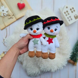 May include: Two handmade crochet snowmen with black hats, orange carrot noses, and red and green scarves. One snowman has a green star, the other a red star. The snowmen have brown feet and are on a fluffy white surface.