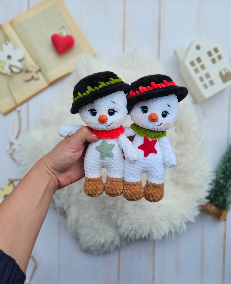 May include: Two handmade crochet snowmen with black hats, orange carrot noses, and red and green scarves. One snowman has a green star, the other a red star. The snowmen have brown feet and are on a fluffy white surface.