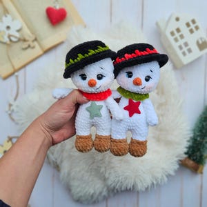 May include: Two handmade crochet snowmen with black hats, orange carrot noses, and red and green scarves. One snowman has a green star, the other a red star. The snowmen have brown feet and are on a fluffy white surface.