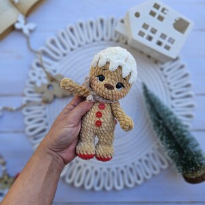 May include: A crocheted gingerbread man doll with a white icing-like top, a tan body, and red accents on the feet and buttons. The doll has a small scarf and is held in a hand. The doll is a festive holiday decoration.