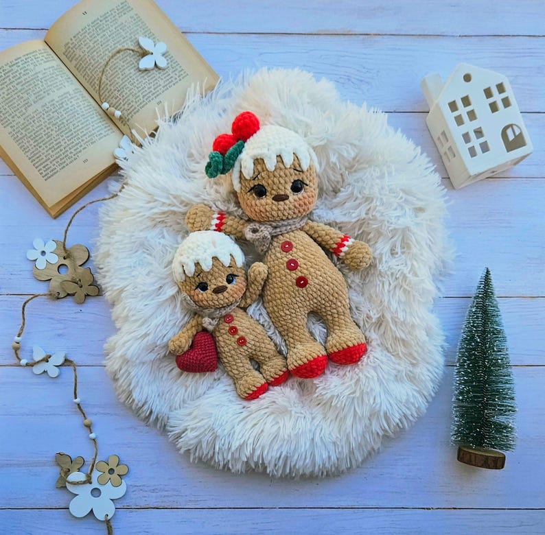 May include: Two crocheted gingerbread man dolls with white icing, red and white striped arms, and red feet, resting on a fluffy white surface. One doll holds a red heart. A small Christmas tree and a white house-shaped decoration are also present.