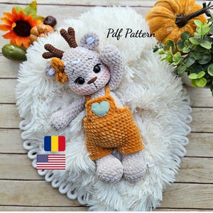 Könnte beinhalten: Gehäkeltes Rentier-Plüschtier mit orangefarbener Latzhose und Herzdetail, liegend auf einer flauschigen weißen Oberfläche. Das Rentier hat braune Geweihe und einen hellgrauen Körper. Der Text "Pdf Pattern" ist sichtbar.
