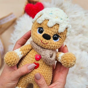 May include: A handmade, crocheted gingerbread man doll with a white icing design and sprinkles. The doll has a tan body, a brown scarf, and a red heart-shaped button. The doll is being held in someone's hands.