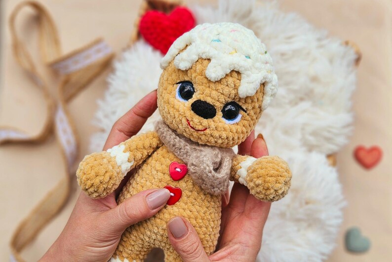 May include: A handmade, crocheted gingerbread man doll with a white icing design and sprinkles. The doll has a tan body, a brown scarf, and a red heart-shaped button. The doll is being held in someone's hands.