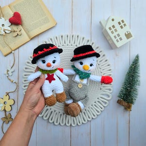 May include: Two handmade crochet snowmen. Each snowman has a black hat with red trim, a carrot nose, and a green scarf. One snowman wears a red star, the other wears gray overalls. They are on a white, textured surface.