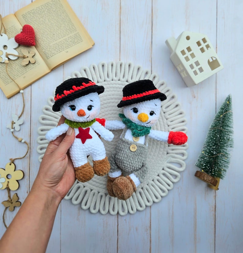 May include: Two handmade crochet snowmen. Each snowman has a black hat with red trim, a carrot nose, and a green scarf. One snowman wears a red star, the other wears gray overalls. They are on a white, textured surface.