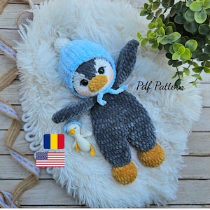 May include: A handmade crochet penguin doll in gray, white, and yellow, wearing a light blue hat. The doll is lying on a fluffy white surface. A smaller penguin doll is also present. The text "Pdf Pattern" is visible.