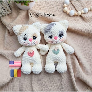 May include: Two handmade white crochet cat dolls with patches of tan and gray on their heads. Each cat has a pink nose, black eyes, and a strawberry or flower applique on the chest. The text "Pdf Pattern" is visible.