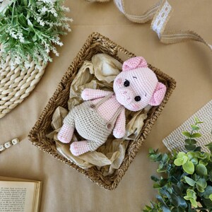 May include: A crocheted pink pig wearing a beige crocheted outfit with overalls. The pig is lying in a wicker basket on a brown surface.