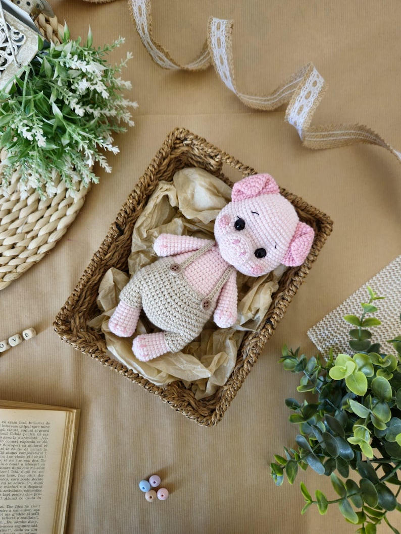 May include: A crocheted pink pig wearing a beige crocheted outfit with overalls. The pig is lying in a wicker basket on a brown surface.