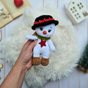 May include: A handmade snowman doll with a black hat trimmed with red, a green scarf, and a red star on its chest. The snowman has brown boots and a carrot nose. The doll is made of white yarn and is a festive holiday decoration.