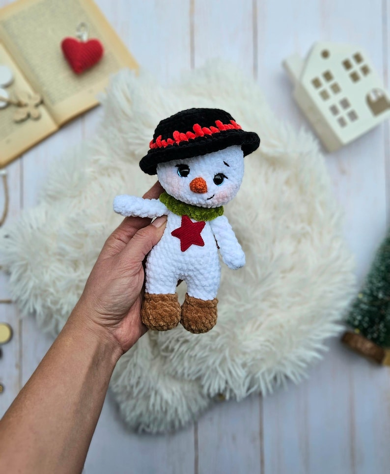 May include: A handmade snowman doll with a black hat trimmed with red, a green scarf, and a red star on its chest. The snowman has brown boots and a carrot nose. The doll is made of white yarn and is a festive holiday decoration.