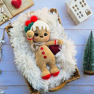 May include: A handmade crochet gingerbread man doll with red and green accents, lying in a woven basket on a fluffy white surface. The doll has a beige body, red buttons, and red feet. A small red heart and a miniature Christmas tree are also in the image.