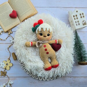 May include: A handmade crochet gingerbread man doll with a red heart, red buttons, and a scarf. The doll has a white frosting-like hair and is lying on a fluffy white surface. A small Christmas tree and a decorative house are in the background.