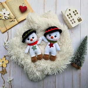 May include: Two handmade crochet snowmen with black hats, red and green scarves, and star accents. The snowmen have carrot noses and brown feet. They are displayed on a fluffy white surface with a small Christmas tree and decorative elements.