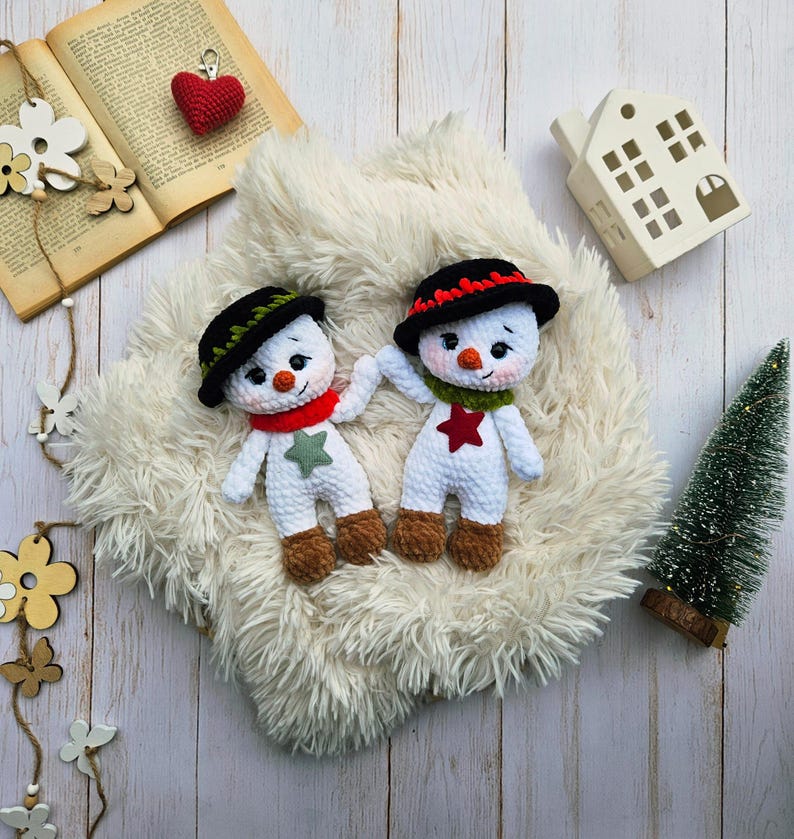 May include: Two handmade crochet snowmen with black hats, red and green scarves, and star accents. The snowmen have carrot noses and brown feet. They are displayed on a fluffy white surface with a small Christmas tree and decorative elements.