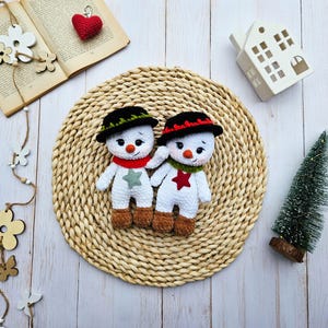 May include: Two handmade crochet snowmen with black hats, red and green scarves, and star accents. The snowmen have brown feet and carrot noses. They are positioned on a woven mat, with a small Christmas tree and a decorative house in the background.