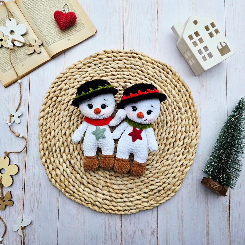 May include: Two handmade crochet snowmen with black hats, red and green scarves, and star accents. The snowmen have brown feet and carrot noses. They are positioned on a woven mat, with a small Christmas tree and a decorative house in the background.