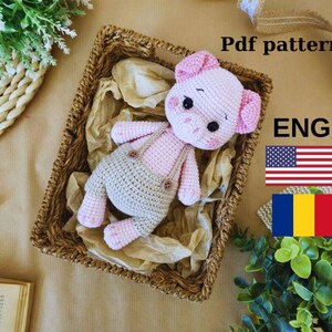 May include: A crocheted pink pig amigurumi toy with a beige crocheted outfit and a brown wicker basket. The image includes the text "Pdf pattern ENG" and flags for the United States and Romania.