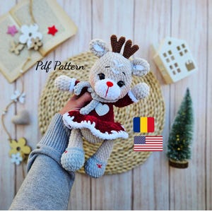 May include: A crocheted reindeer doll with a red dress, white trim, and a heart-shaped detail. The doll has brown antlers, a red nose, and gray shoes with red star accents. The text "Pdf Pattern" is visible.