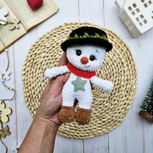 May include: A handmade crochet snowman doll with a black hat, red scarf, and a green star on its front. The snowman has a carrot nose and brown boots. The doll is white with black eyes and is approximately 5 inches tall.