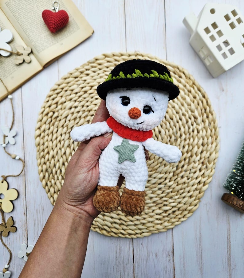 May include: A handmade crochet snowman doll with a black hat, red scarf, and a green star on its front. The snowman has a carrot nose and brown boots. The doll is white with black eyes and is approximately 5 inches tall.