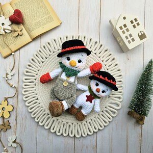 May include: Two handmade crochet snowmen. One snowman wears a black hat with a red band, a green scarf, red mittens, and gray overalls. The other snowman has a black hat with a red band, a green scarf, and a red star on its chest.
