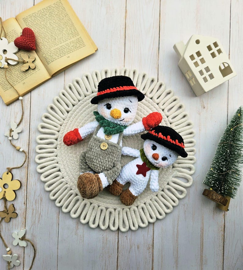 May include: Two handmade crochet snowmen. One snowman wears a black hat with a red band, a green scarf, red mittens, and gray overalls. The other snowman has a black hat with a red band, a green scarf, and a red star on its chest.