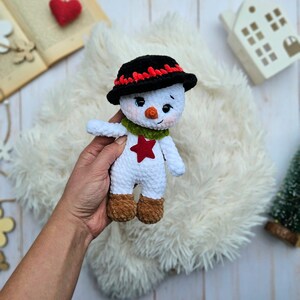 May include: A handmade crochet snowman doll. The snowman has a black hat with red trim, a green scarf, and a red star on its chest. It has brown boots and is holding its arms out. The doll is white with a cute face.