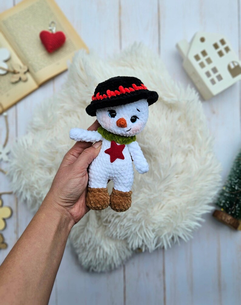 May include: A handmade crochet snowman doll. The snowman has a black hat with red trim, a green scarf, and a red star on its chest. It has brown boots and is holding its arms out. The doll is white with a cute face.