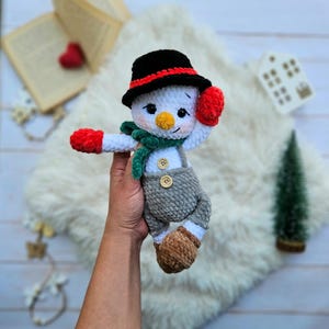 May include: A handmade crochet snowman doll with a black hat, red mittens, and a green scarf. The snowman wears gray overalls with wooden buttons and brown boots. The doll is held in a person's hand against a white fluffy background.