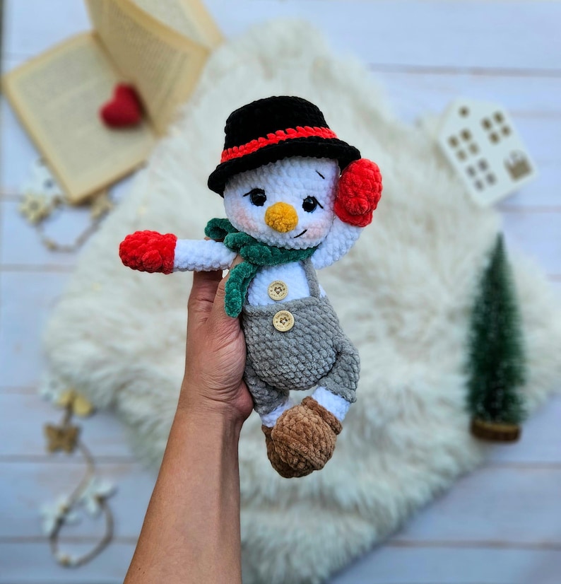 May include: A handmade crochet snowman doll with a black hat, red mittens, and a green scarf. The snowman wears gray overalls with wooden buttons and brown boots. The doll is held in a person's hand against a white fluffy background.